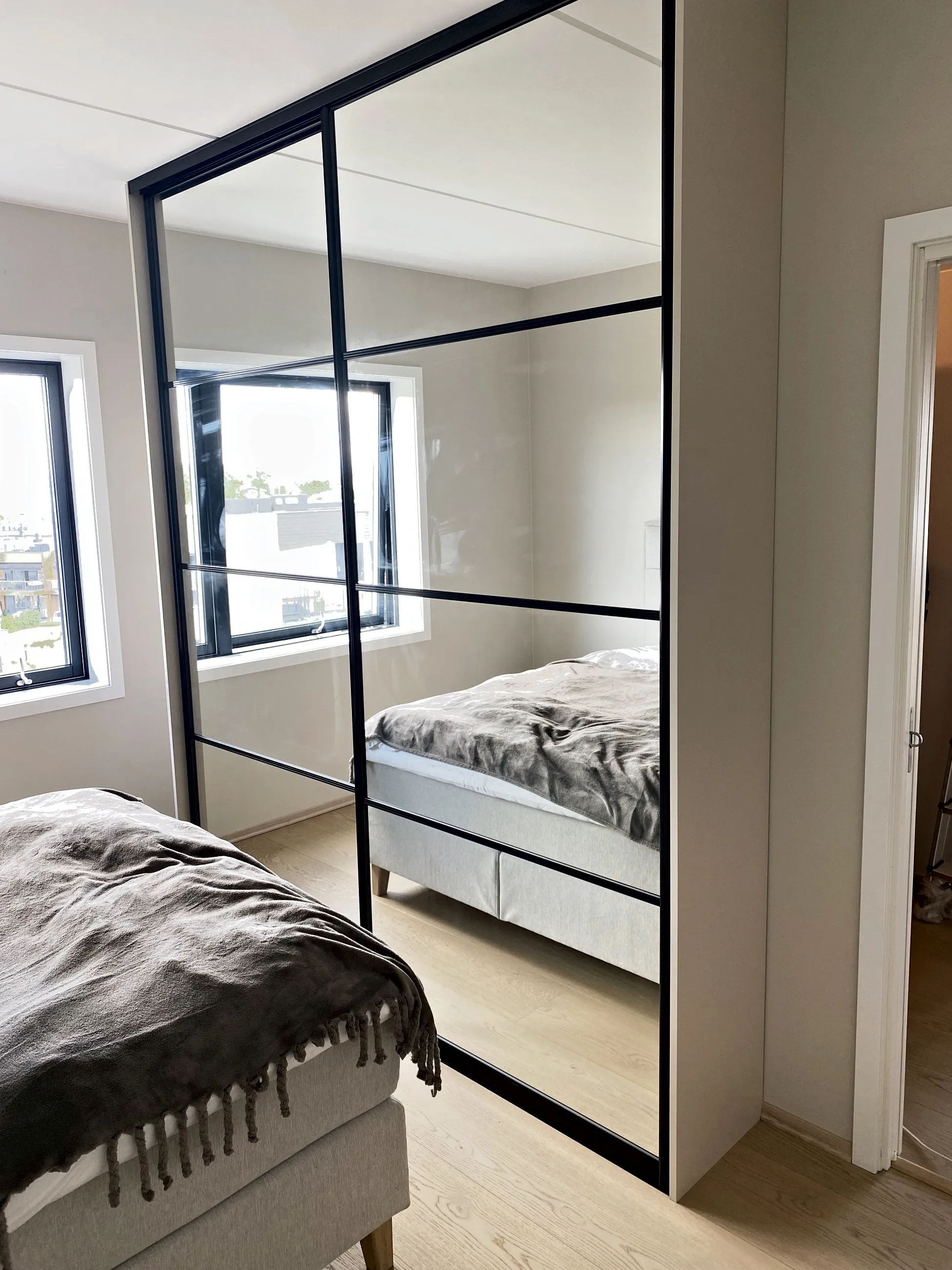 Skyvedører med heldekkende speil i svart ramme, som speiler seng og vindu i et lyst soverom